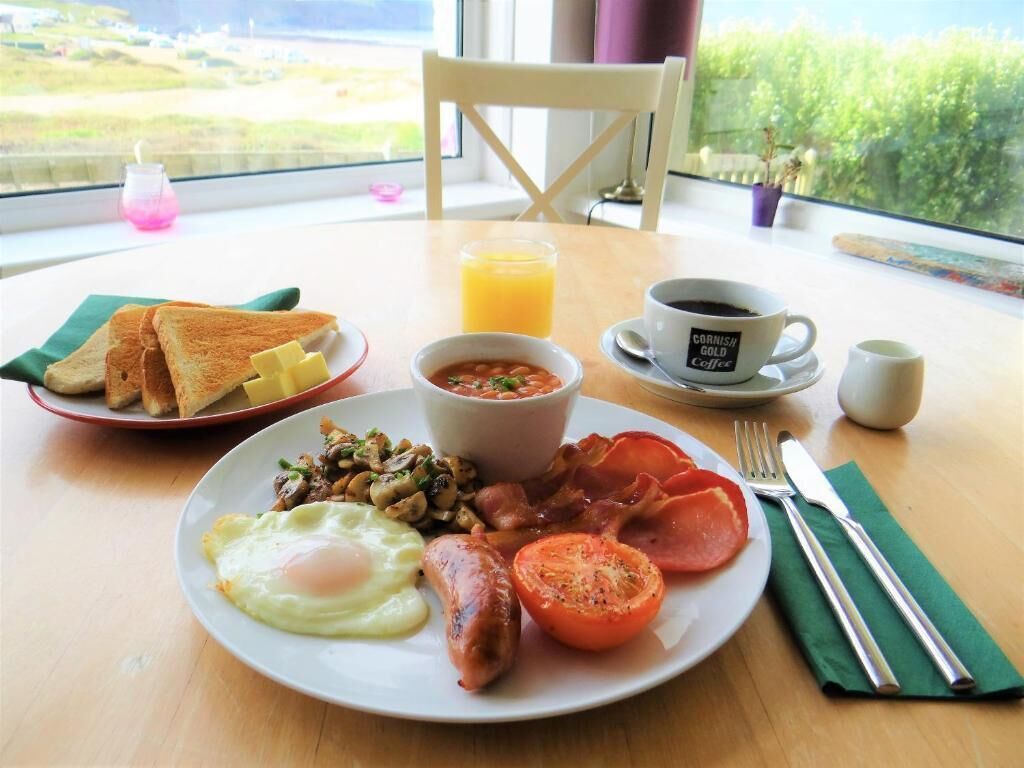 A breakfast plate with toast, eggs, bacon, sausage, and tomato, along with a cup of coffee and a glass of orange juice on a table by a window.