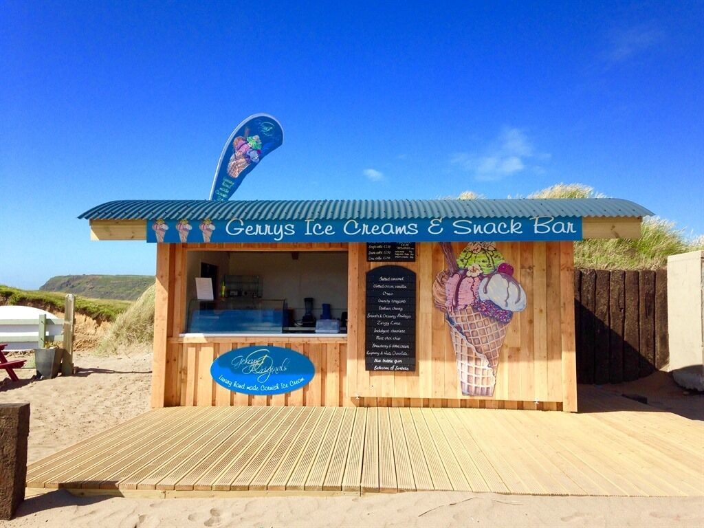 Un bar de helados y snacks frente a la playa llamado 'Gerys Ice Creams & Snack Bar', con estructura de madera, toldo azul y un cartel con la carta.
