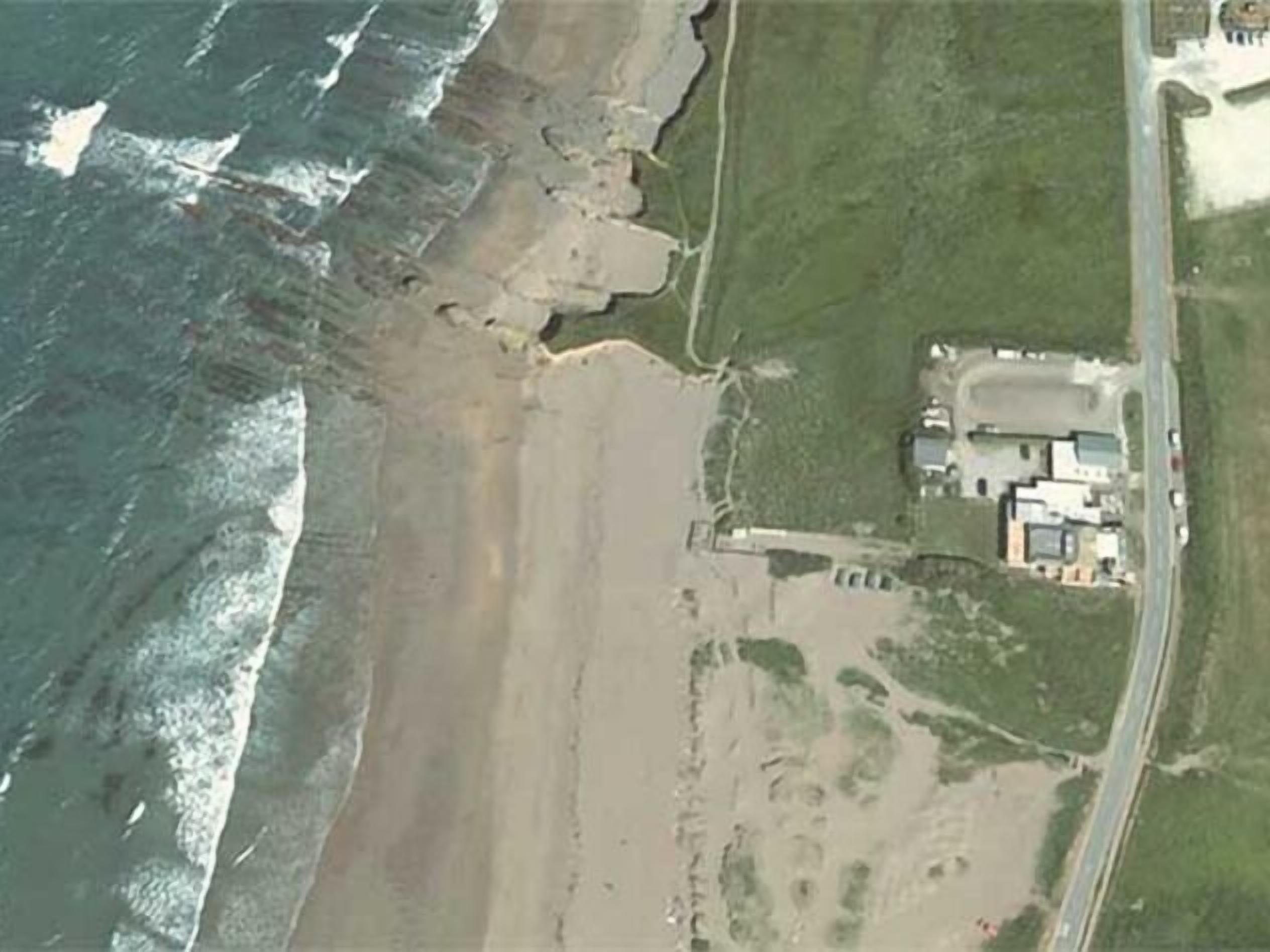 An aerial view of a coastal property with a large beachfront, a road adjacent to the beach, and a cluster of buildings with a central structure that appears to be a main building.