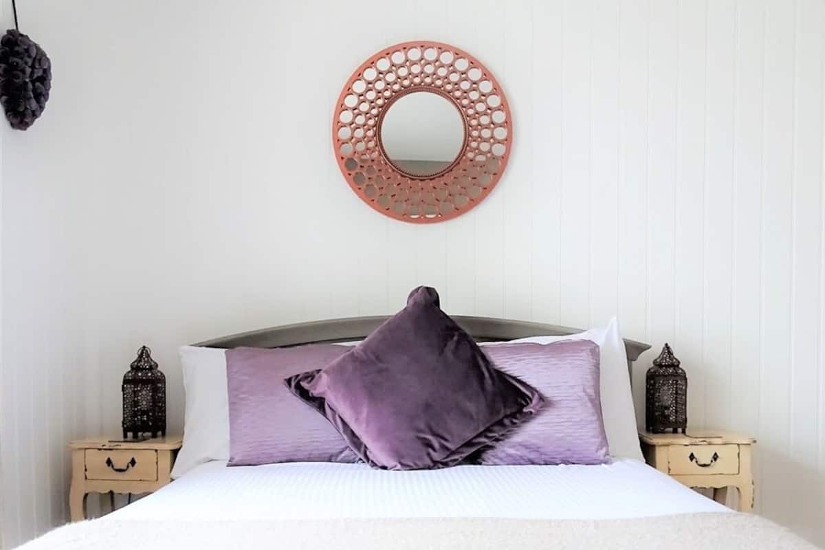 A neatly made bed with a purple pillow, a round mirror on the wall, and two small side tables with decorative lanterns.