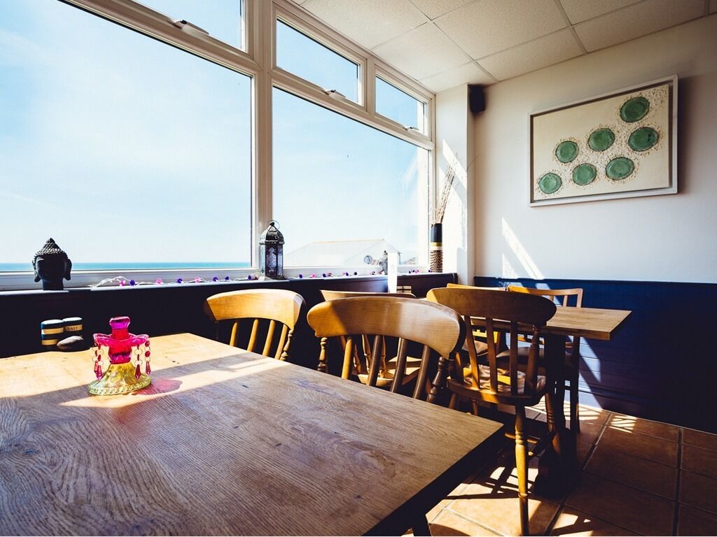 A dining area with wooden tables and chairs, a view of the sea through large windows, and a wall-mounted artwork.