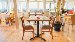 A spacious dining area with wooden tables and chairs, large windows offering a view of the sea, and potted plants adding greenery.