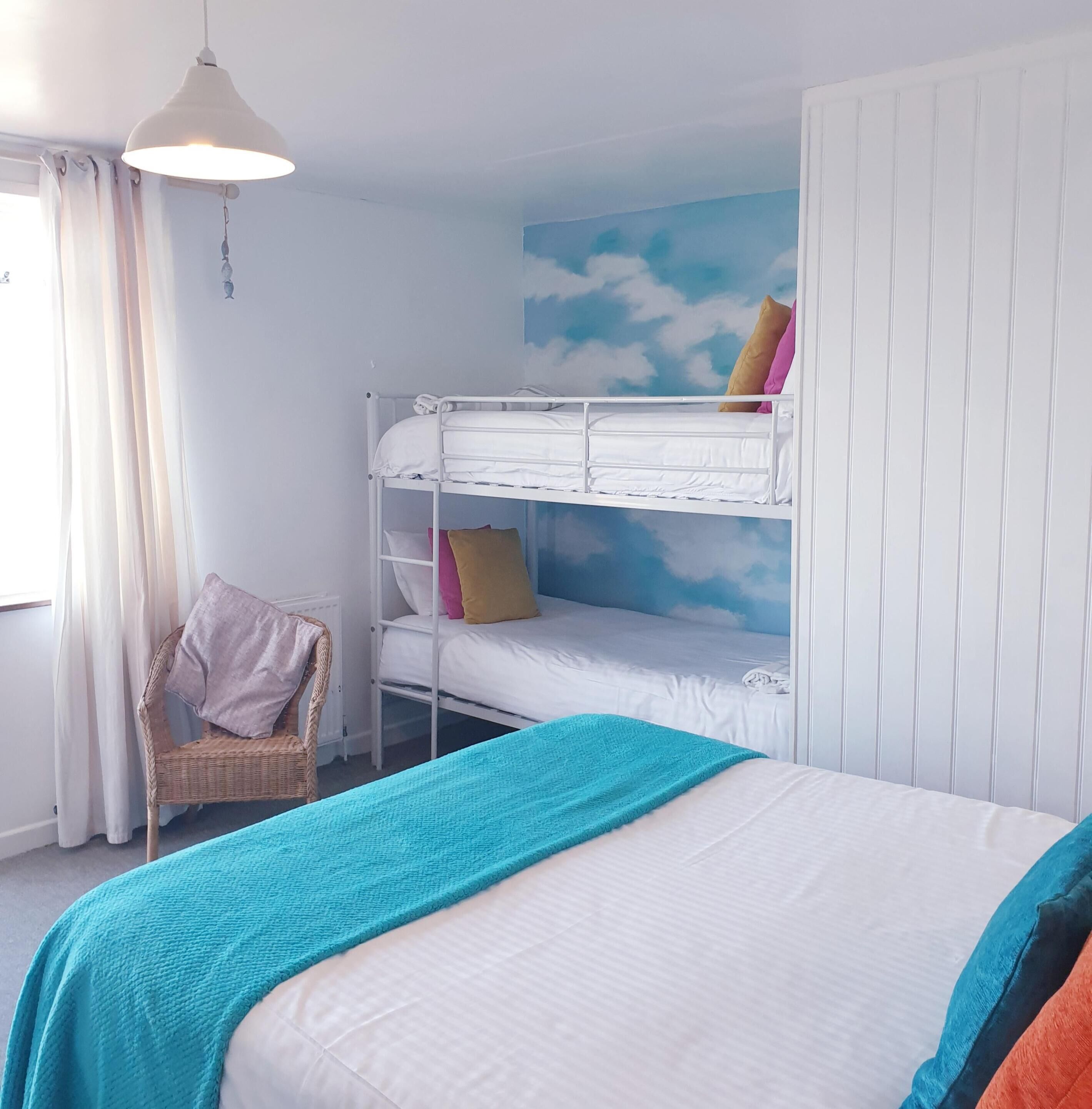A bunk bed room with a white ceiling, a wall mural of clouds, a wicker chair, and a large bed with a teal blanket.