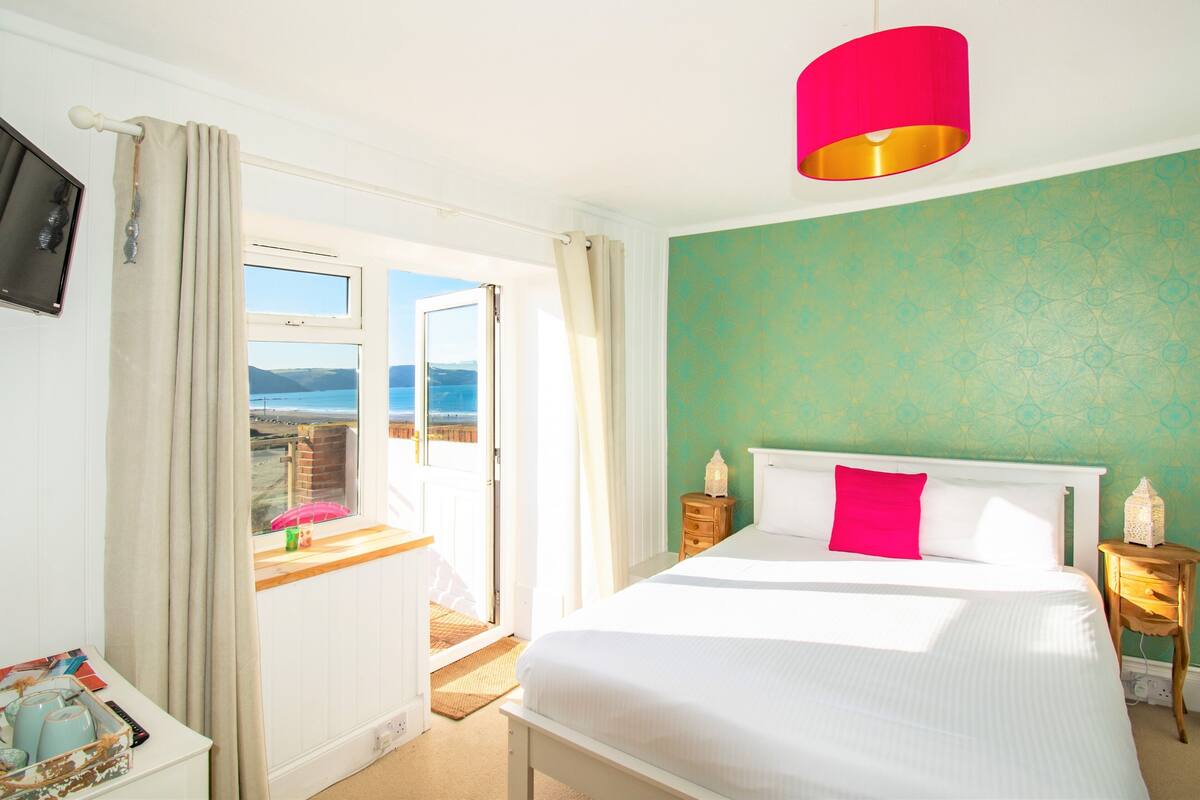 A bedroom with a large bed, a view of the beach, and a teal-colored wall.