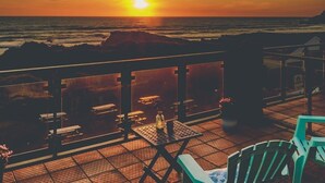 A terrace with a view of the sunset over the ocean, featuring outdoor seating and a railing.