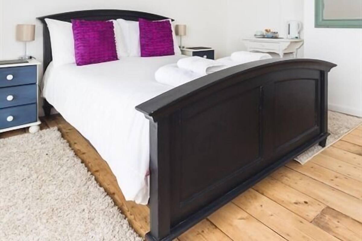 A bedroom with a dark wooden bed, white bedding, and purple pillows. There is a white nightstand with a lamp, a small table with a kettle, and a white dresser.