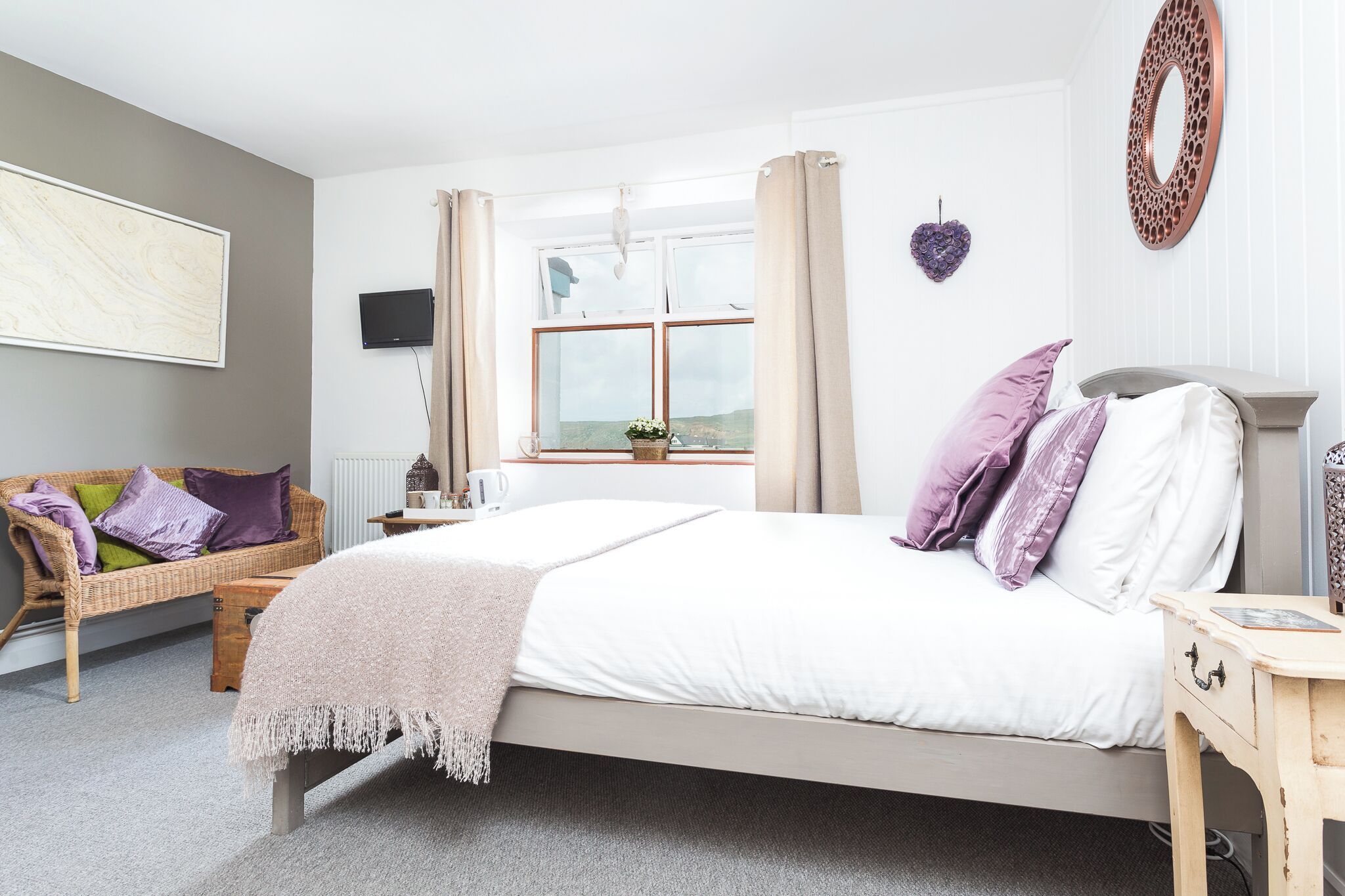 A bedroom with a bed, a bedside table, a nightstand, a window with curtains, a wall-mounted TV, a wicker sofa, and a decorative heart on the wall.