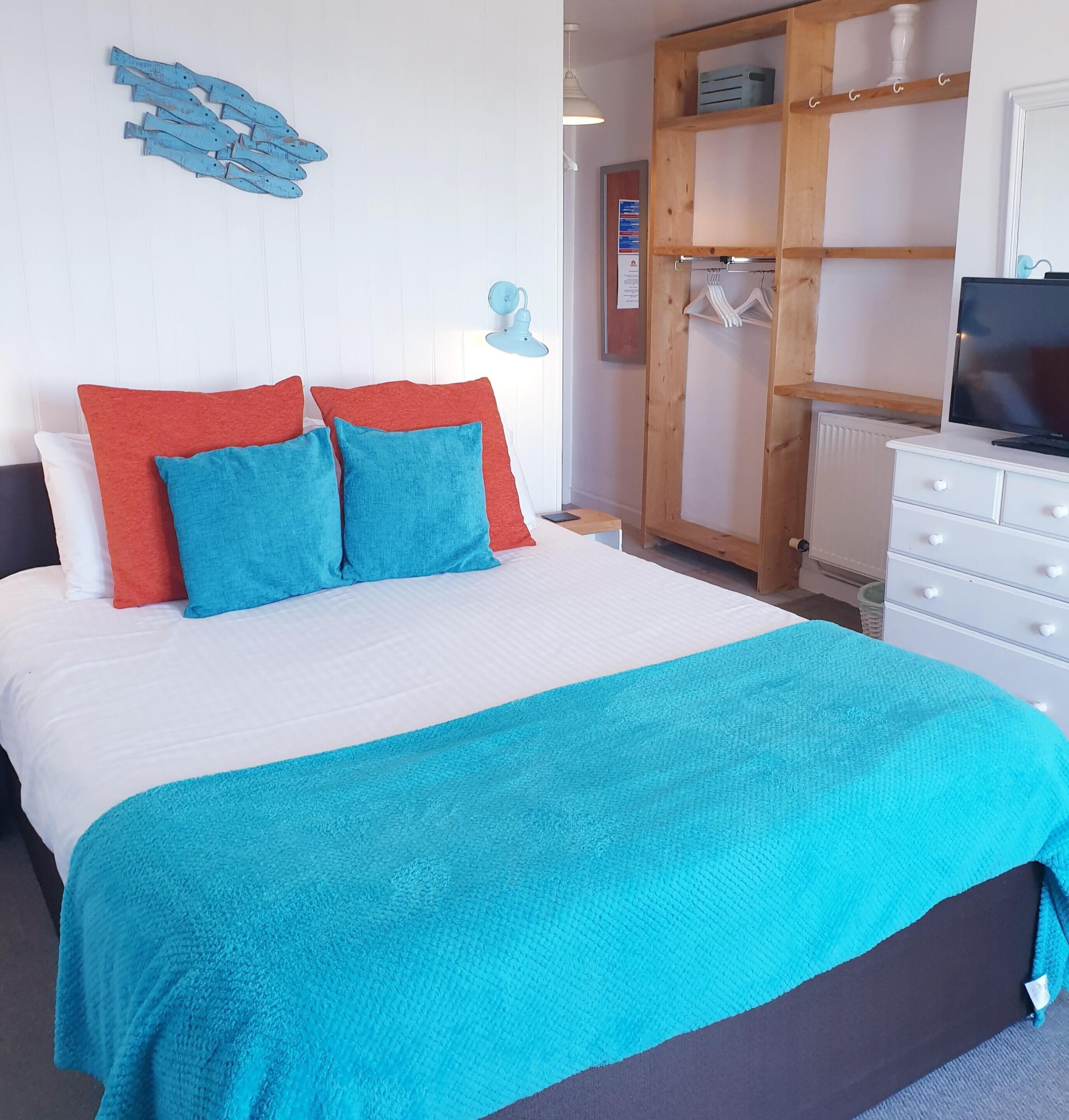 Una cama bien hecha con colcha blanca y azul, dos almohadas rojas y dos azules, un armario de madera con espejo y un televisor sobre una cómoda.