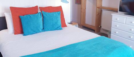 Una cama bien hecha con colcha blanca y azul, dos almohadas rojas y dos azules, un armario de madera con espejo y un televisor sobre una cómoda.