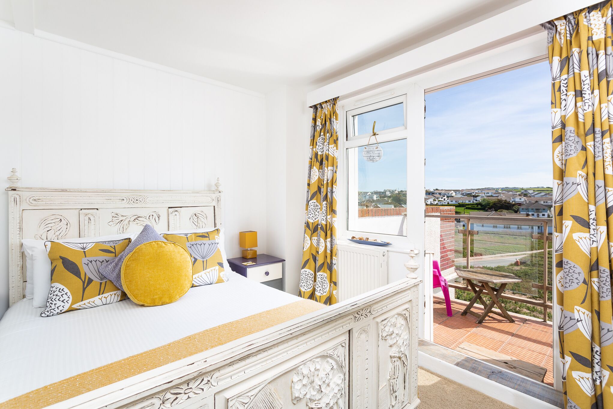 A bedroom with a white bed, yellow and gray patterned pillows, a nightstand with a lamp, and a view of a balcony with a table and chairs.