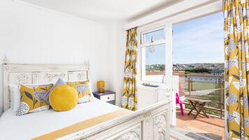 A bedroom with a white bed, yellow and gray patterned pillows, a nightstand with a lamp, and a view of a balcony with a table and chairs.