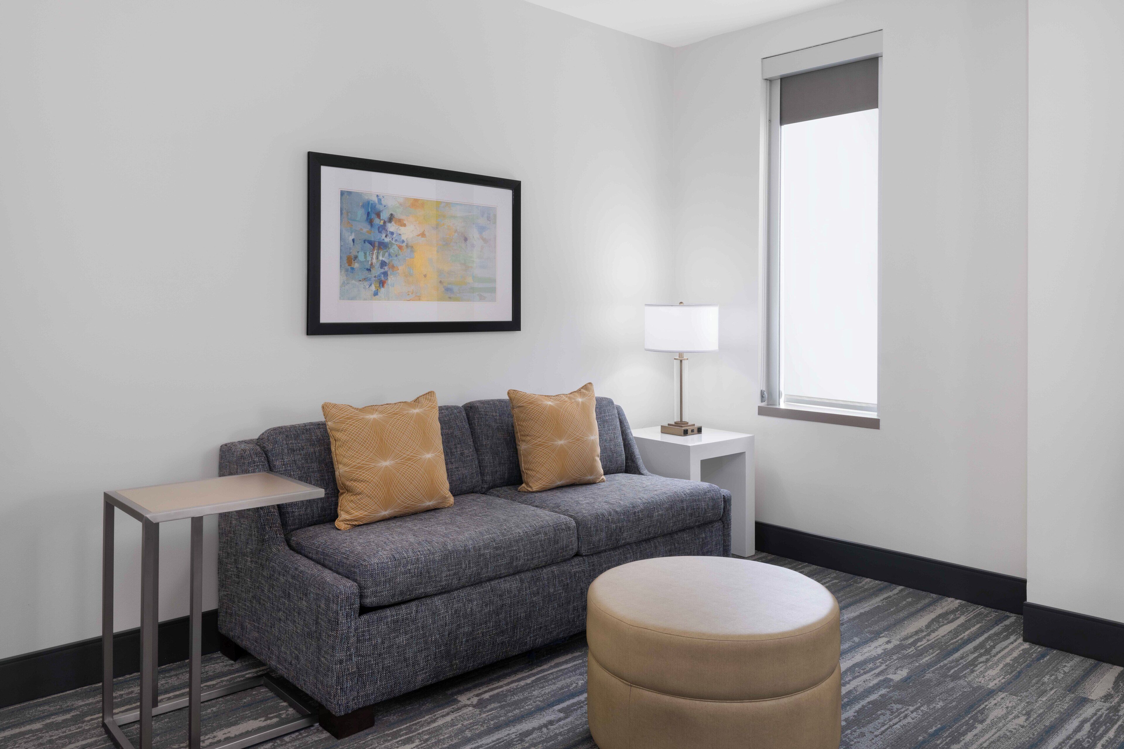 Une chambre moderne avec un canapé gris, deux coussins, une petite table d’appoint avec une lampe et un pouf rond.