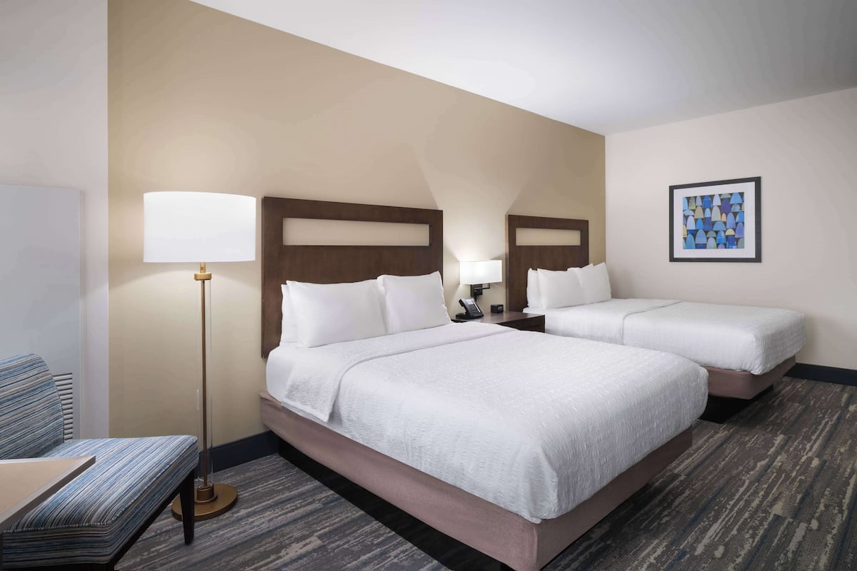 A hotel room with two beds, a bedside table with a lamp, a chair, and a framed picture on the wall.