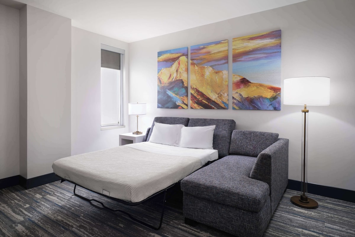 A hotel room with a bed, a gray sofa, a nightstand with a lamp, and three abstract paintings on the wall.