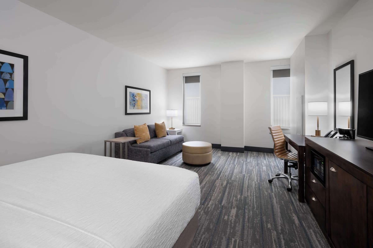A hotel room with a large bed, a gray sofa, a desk with a computer, and a TV.