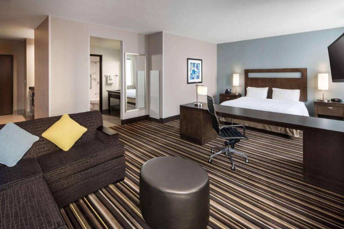 Une chambre d’hôtel comprenant un lit, un bureau, une chaise, un canapé et un pouf rond.