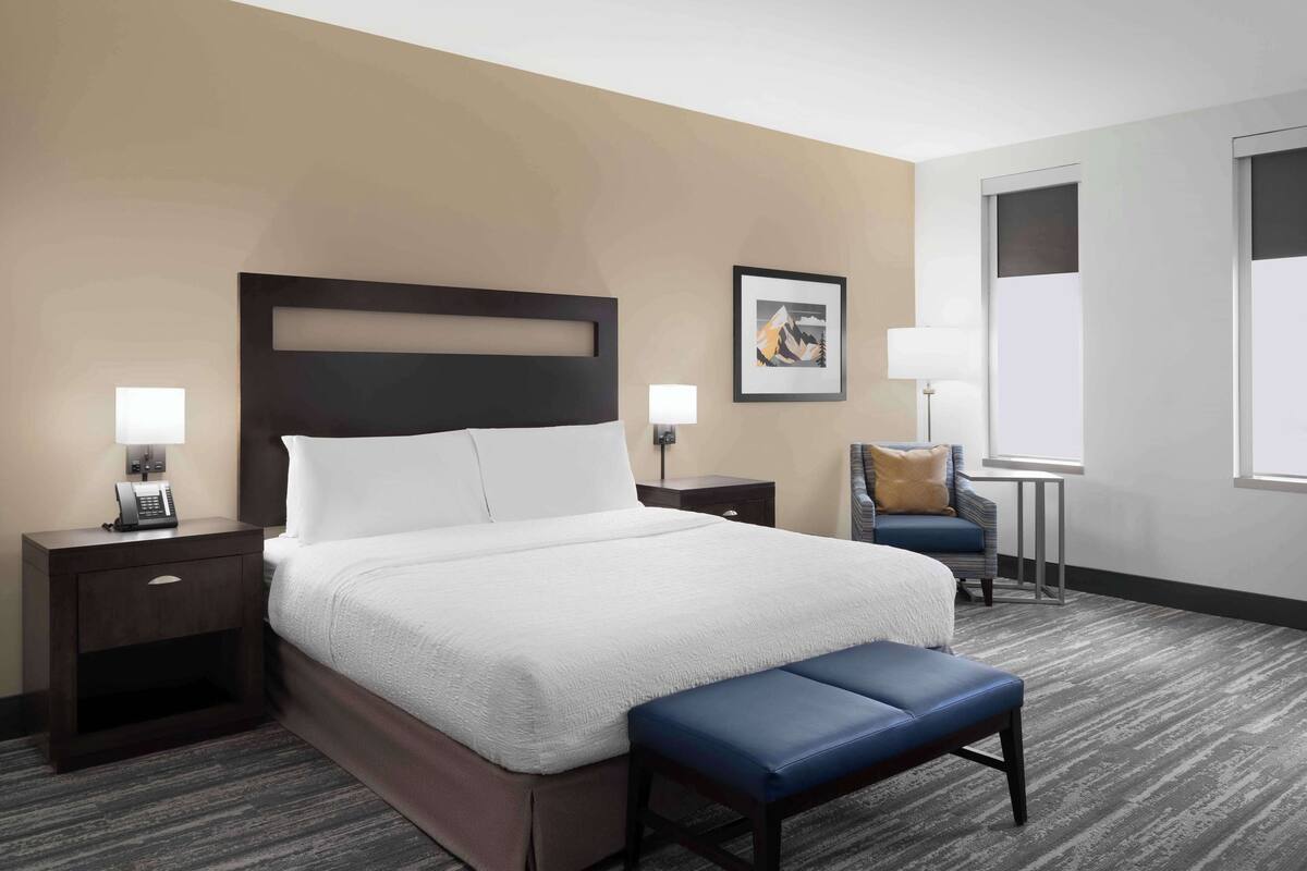 A hotel room with a large bed, two bedside tables with lamps, a chair, a small table, and a window.