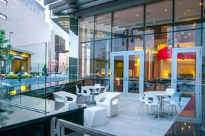 A modern hotel with a glass facade, outdoor seating area, and a visible restaurant inside.
