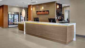 A modern reception area with a wooden reception desk, two computer monitors, a refrigerator stocked with beverages, and a television on a shelf.
