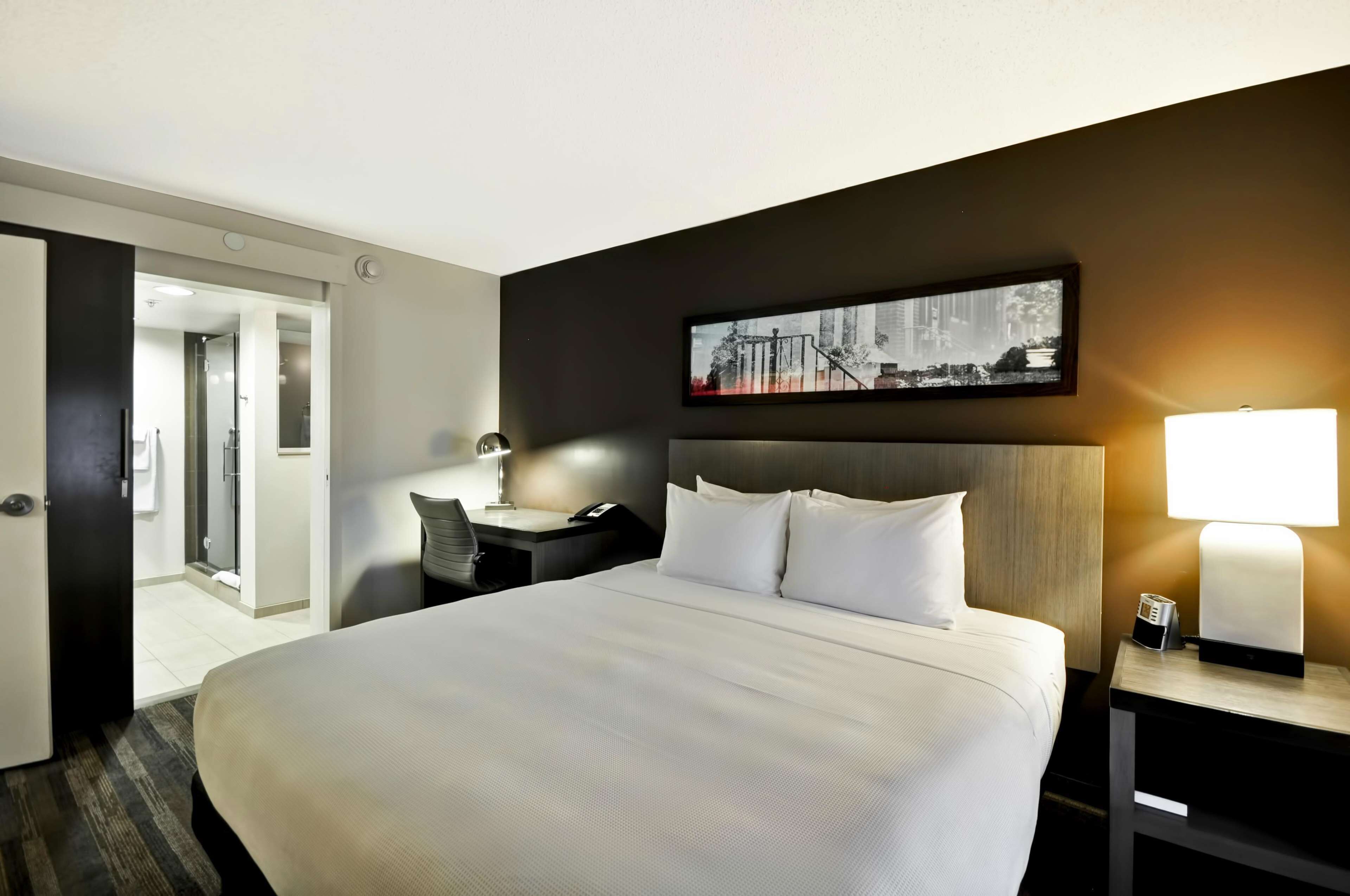 A hotel room with a large bed, a desk with a chair, a nightstand with a lamp, and a framed picture on the wall.