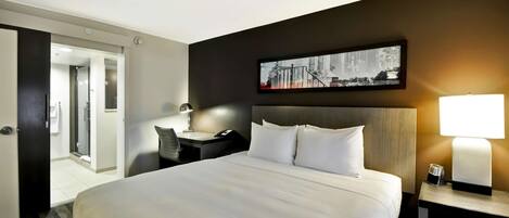 Ein Hotelzimmer mit einem großen Bett, einem Schreibtisch mit Stuhl, einem Nachttisch mit Lampe und einem gerahmten Bild an der Wand.