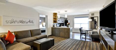 A modern living room with a sofa, coffee table, and a flat-screen TV. There is a kitchen area with a dining table and chairs.