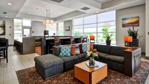 Un hall d’hôtel moderne avec un canapé, un pouf et une table basse. On y trouve également un bar avec des tabourets et une grande fenêtre donnant sur l’extérieur.