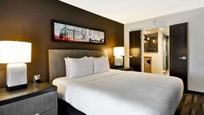 Une chambre d’hôtel avec un grand lit, deux lampes de chevet, une table de chevet avec un téléphone, un tableau encadré au mur et une porte donnant sur une salle de bain.