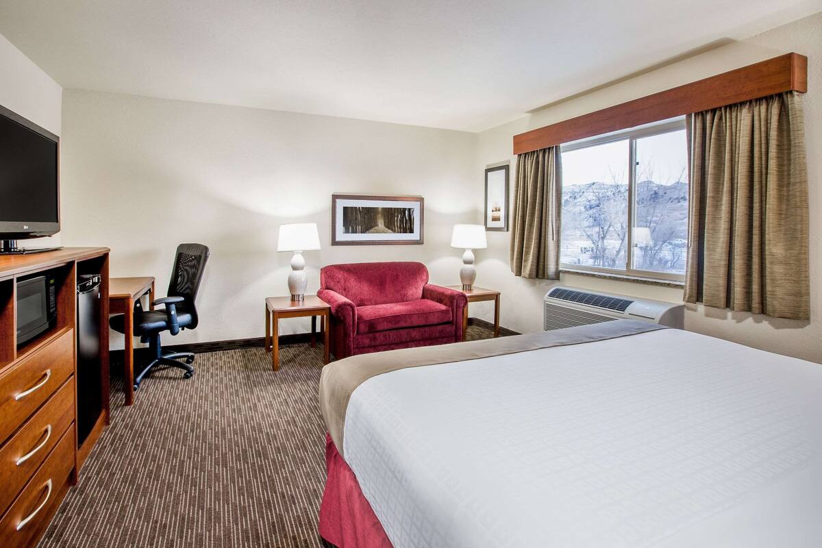 Habitación de hotel con cama, escritorio con computadora, silla, televisor y ventana con cortinas.