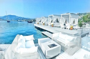 Une piscine à débordement bordée de mobilier d’extérieur blanc et dotée d’un espace salon couvert avec vue sur l’eau.
