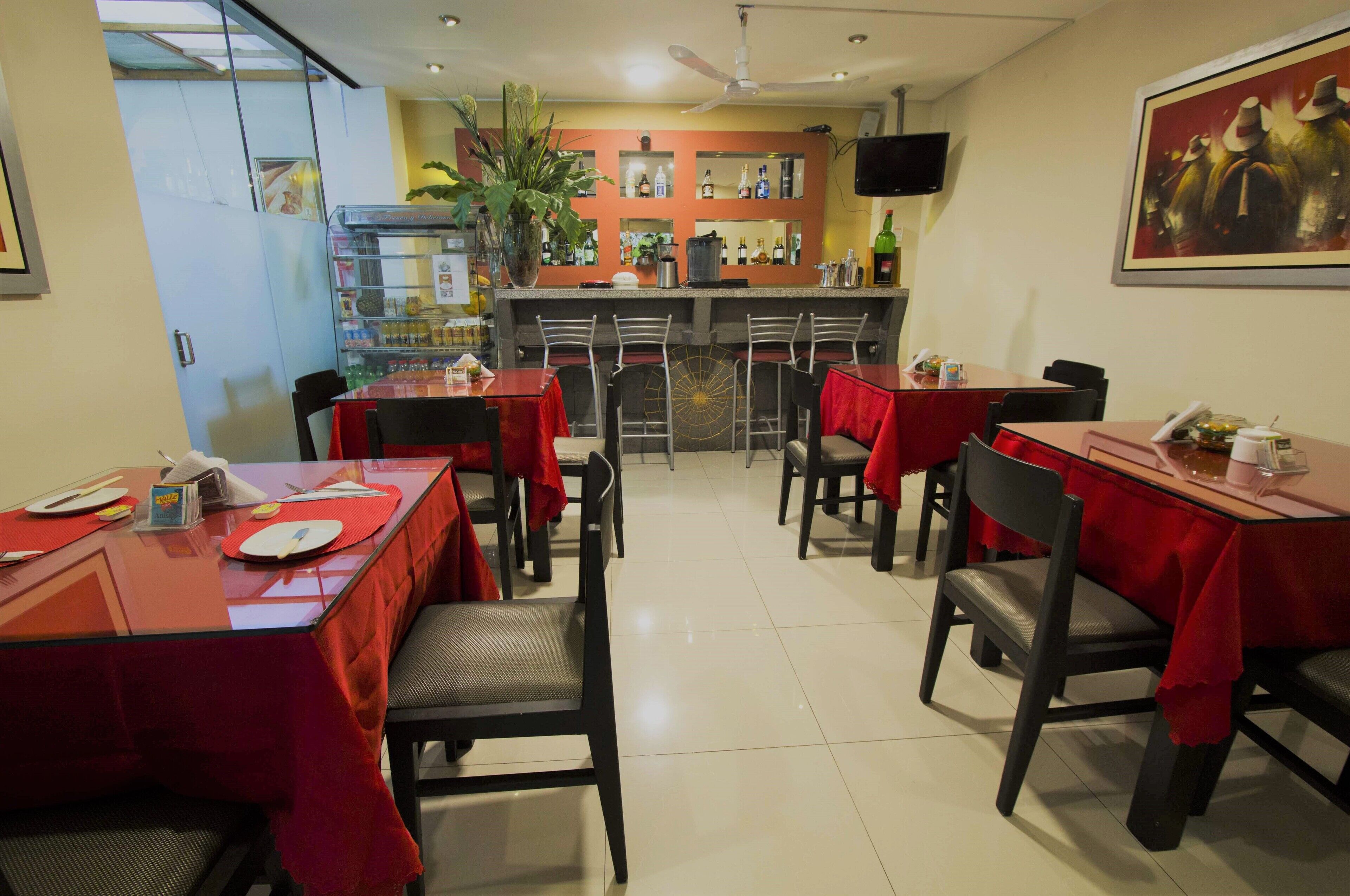 Un restaurante con mesas cubiertas de manteles rojos, sillas, una zona de bar y un televisor en la pared.