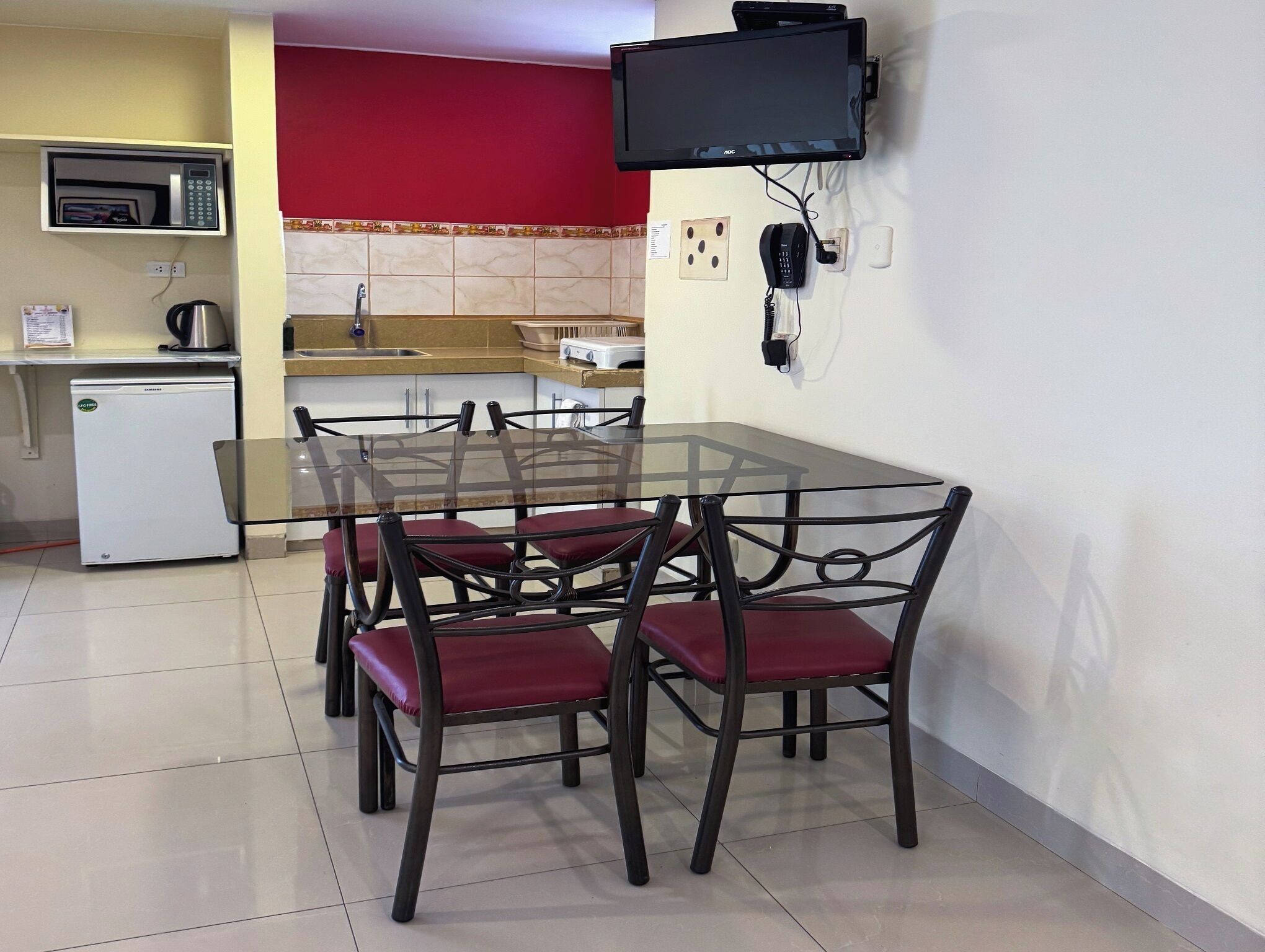 A compact kitchen with a microwave, a small refrigerator, a sink, and a dining area with a glass table and red chairs.