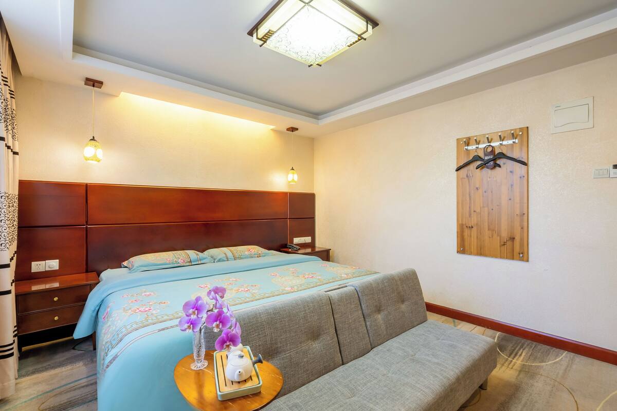 A hotel room with a double bed, a sofa, a wooden headboard, a wall-mounted coat rack, and a small table with a teapot and orchid.