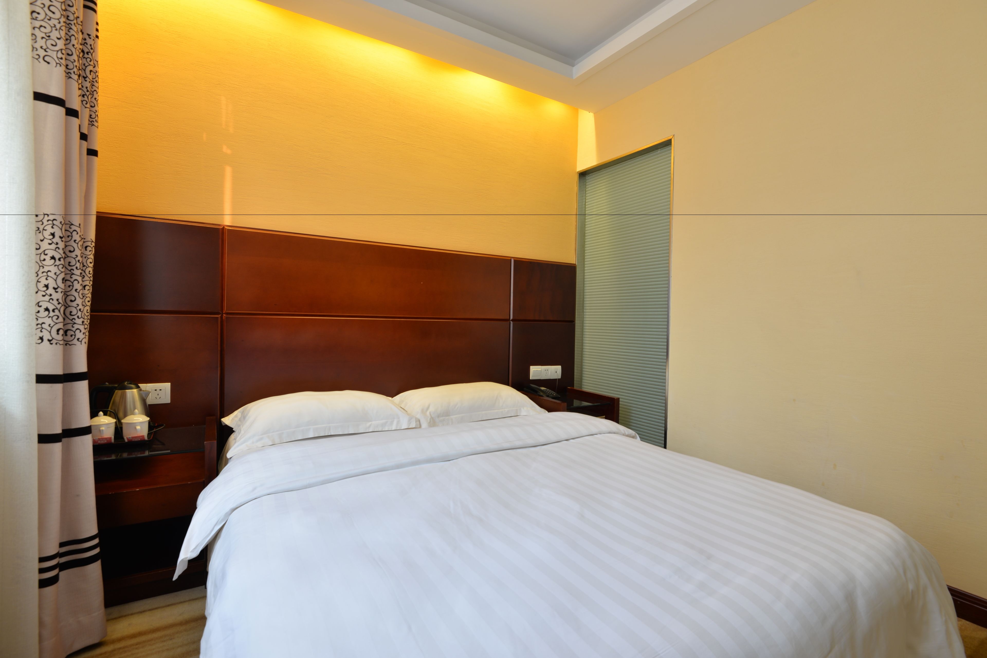 A hotel room with a large bed, a nightstand with a lamp, a power outlet, and a window with blinds.