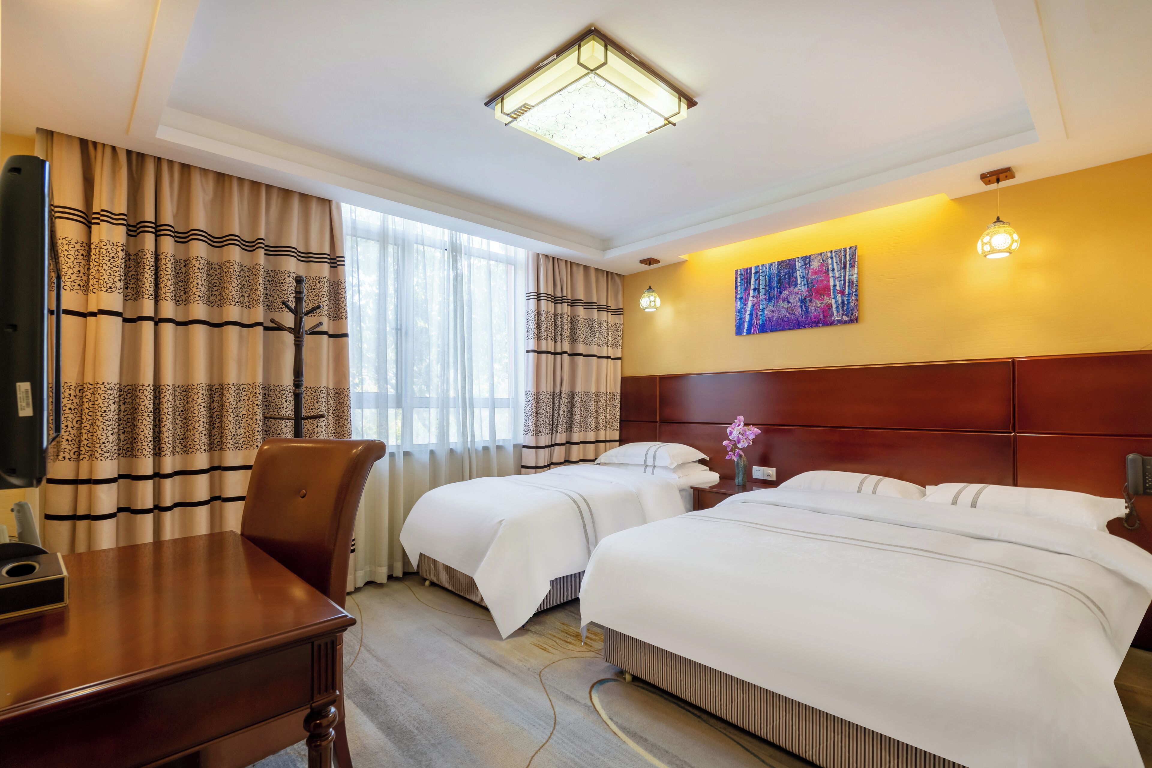 A hotel room with two beds, a desk, a chair, a painting on the wall, and a ceiling light.