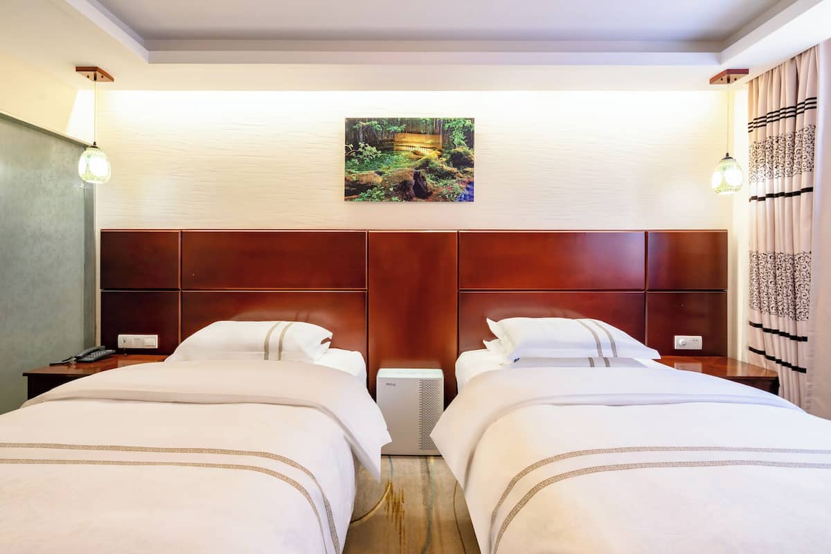 A hotel room with two beds, a wooden headboard, a painting of a forest scene, and a nightstand with a lamp.