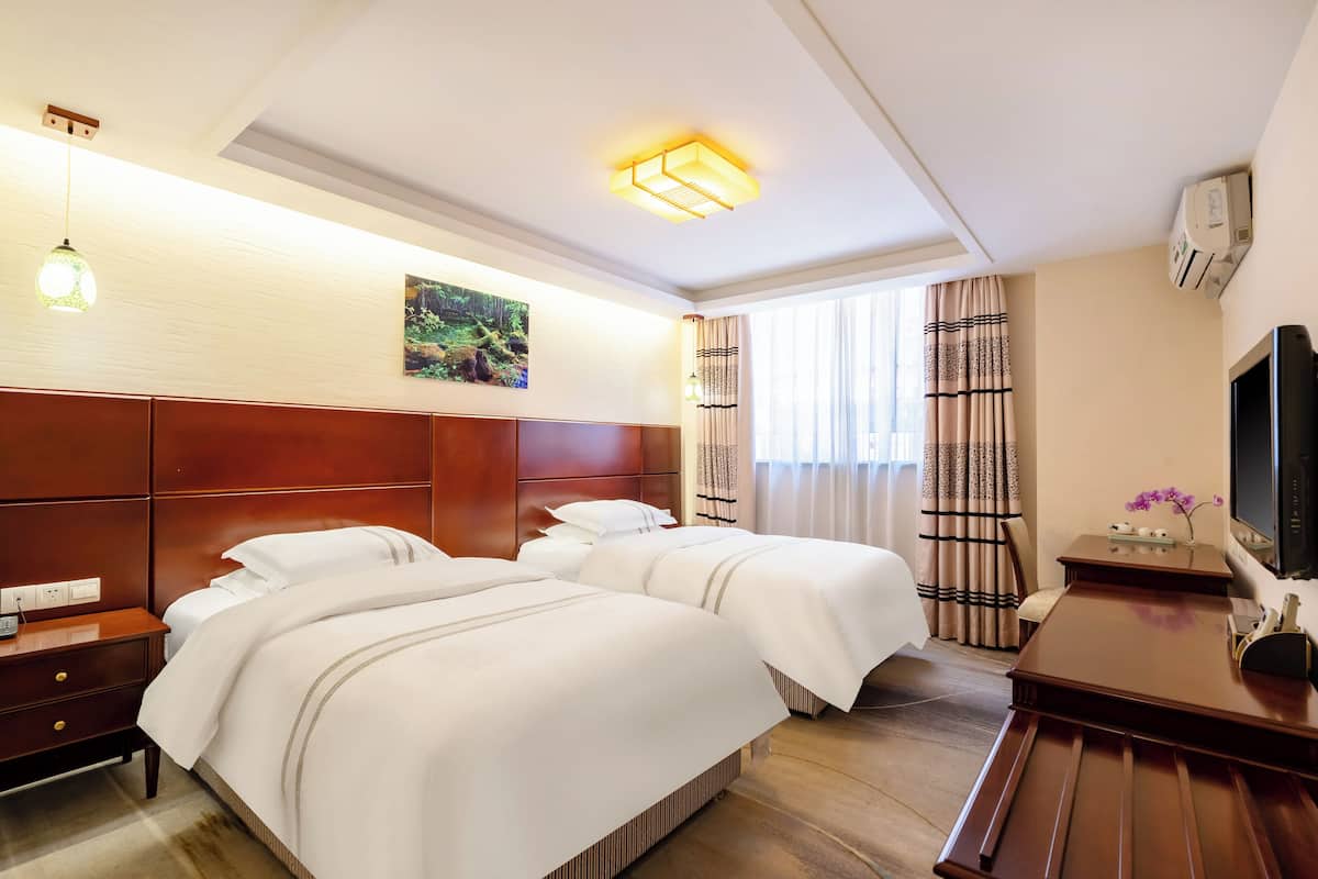 A hotel room with two beds, a wooden headboard, a nightstand, a TV, and a window with curtains.