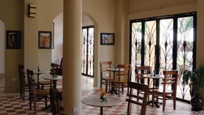 A spacious indoor area with multiple wooden tables and chairs, large windows with decorative iron grilles, and a ceiling fan. - The Columns (Kampot)