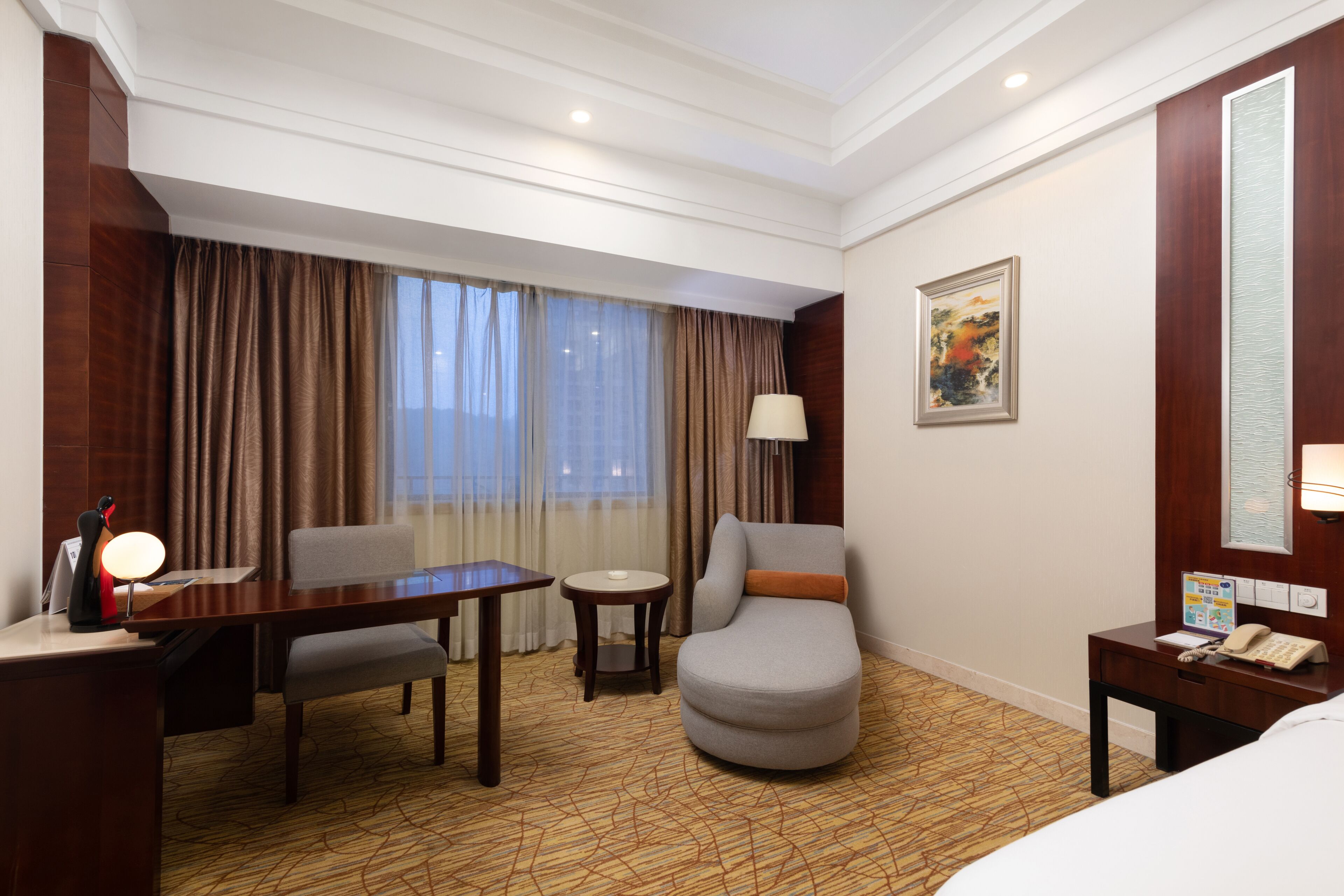 A hotel room with a desk, chair, armchair, bedside table, lamp, and a painting on the wall.