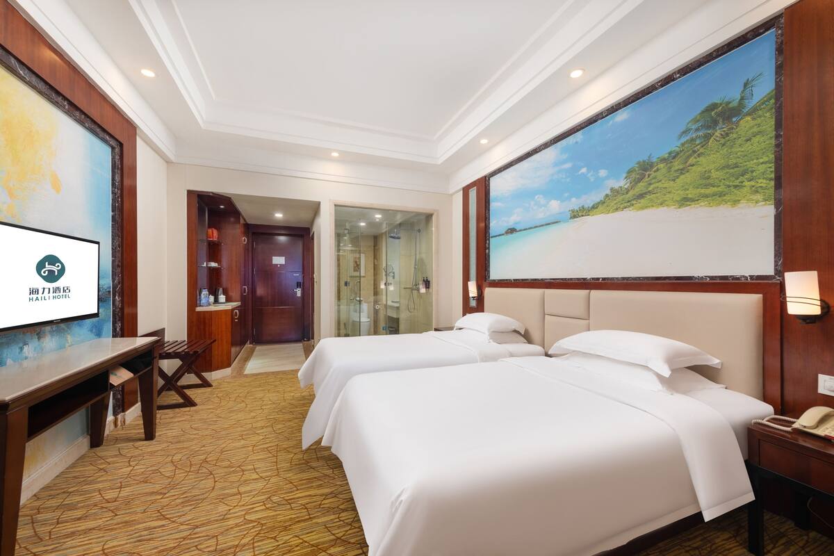 A hotel room with two beds, a desk, a chair, a bathroom with a glass door, and a large beach mural on the wall.