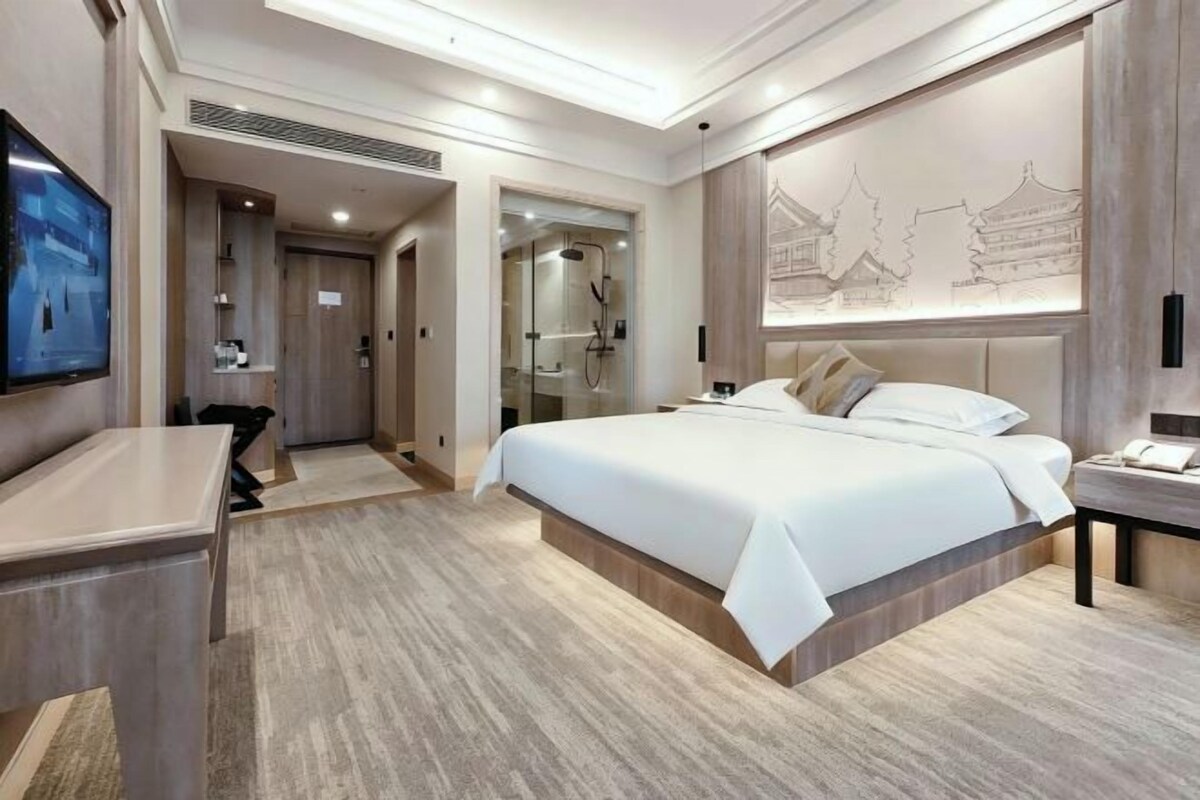 Habitación de hotel moderna con una cama grande, un escritorio, una televisión y un baño con puerta de vidrio.