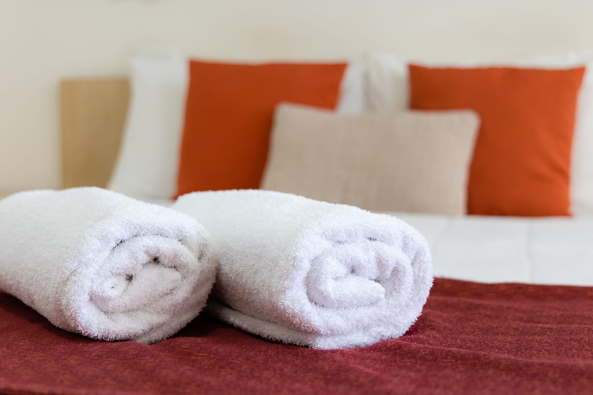 A neatly made white towel on a red bedspread, with a white bed and orange pillows in the background.