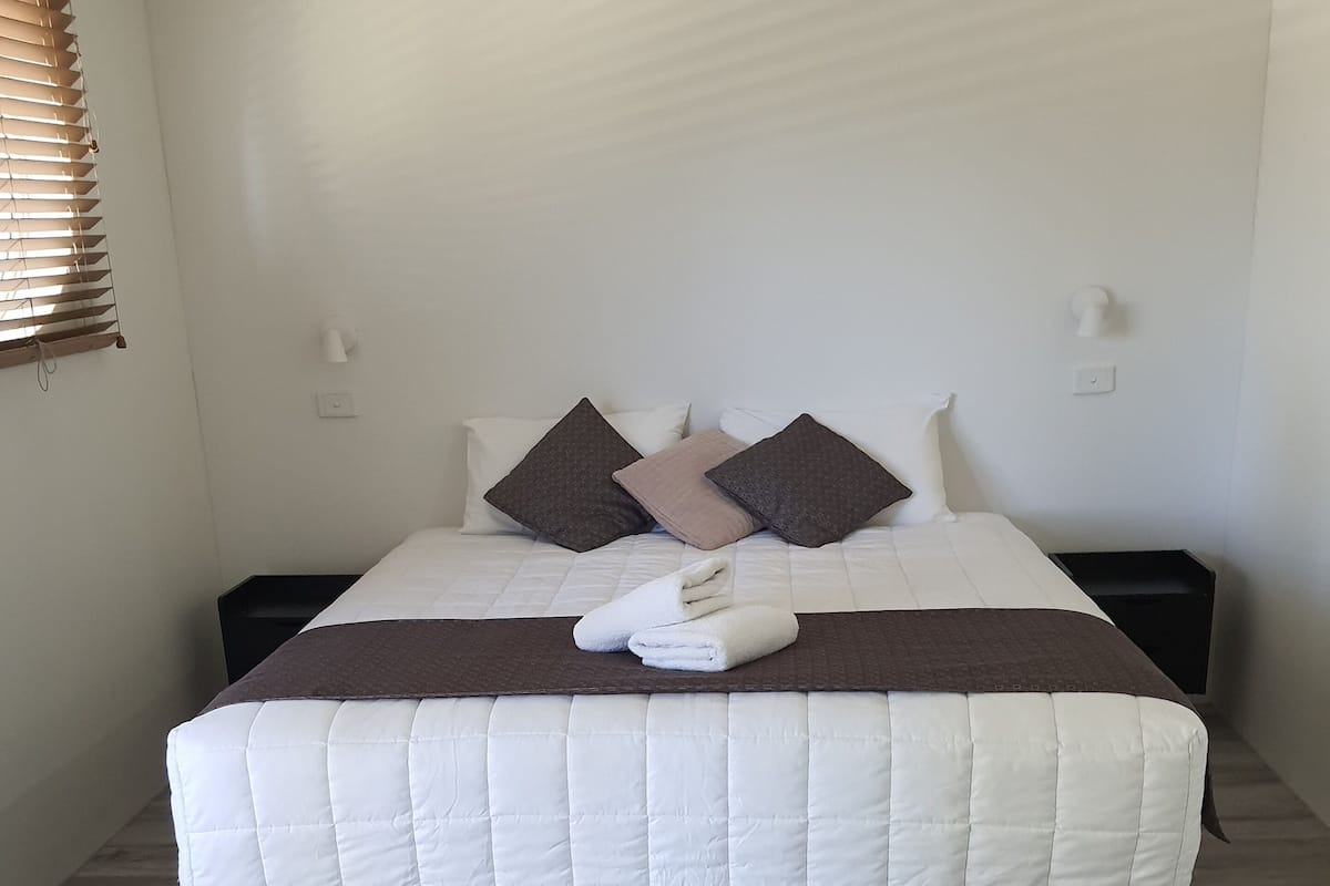 A neatly made bed with a white and brown bedspread, two dark pillows, and a folded towel on a dark headboard.