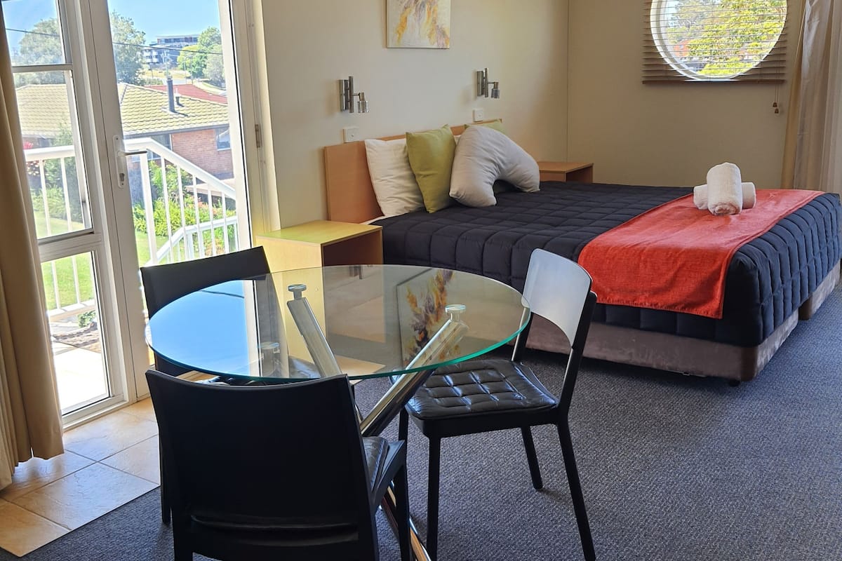 A hotel room with a bed, a glass table with chairs, a ceiling fan, and a view of the outside.
