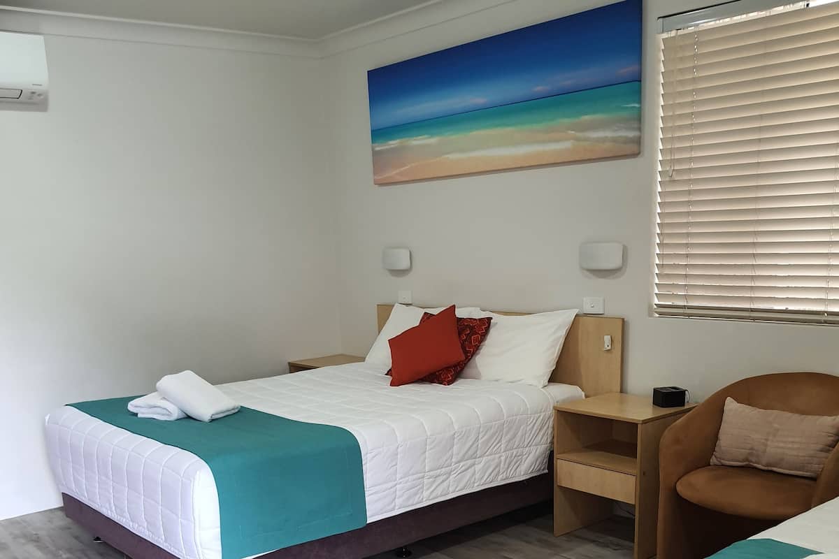 A hotel room with two beds, a wooden floor, a painting of a beach, and a window with blinds.