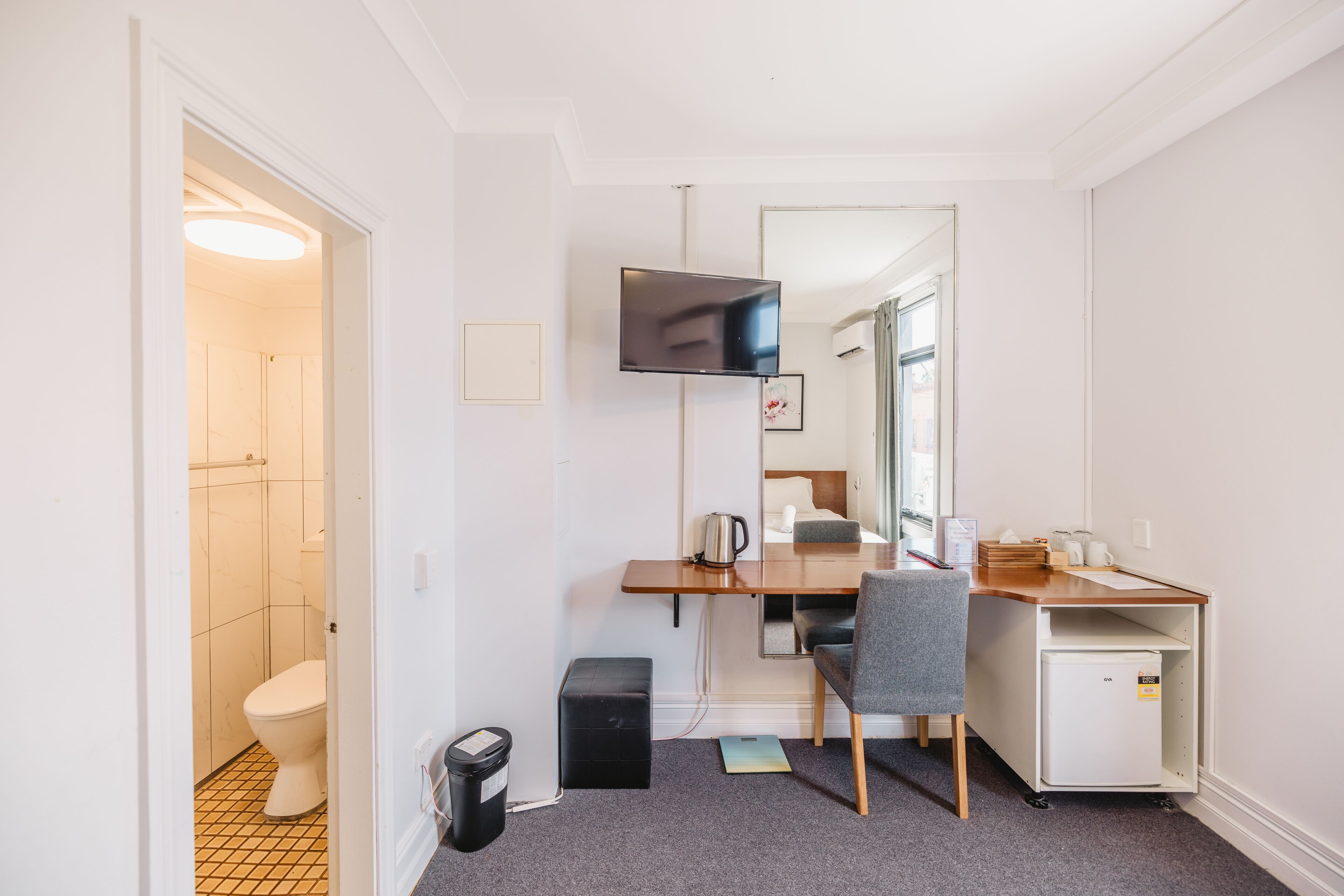 Habitación de hotel compacta con escritorio de madera, una silla, un refrigerador pequeño, televisor y espejo.