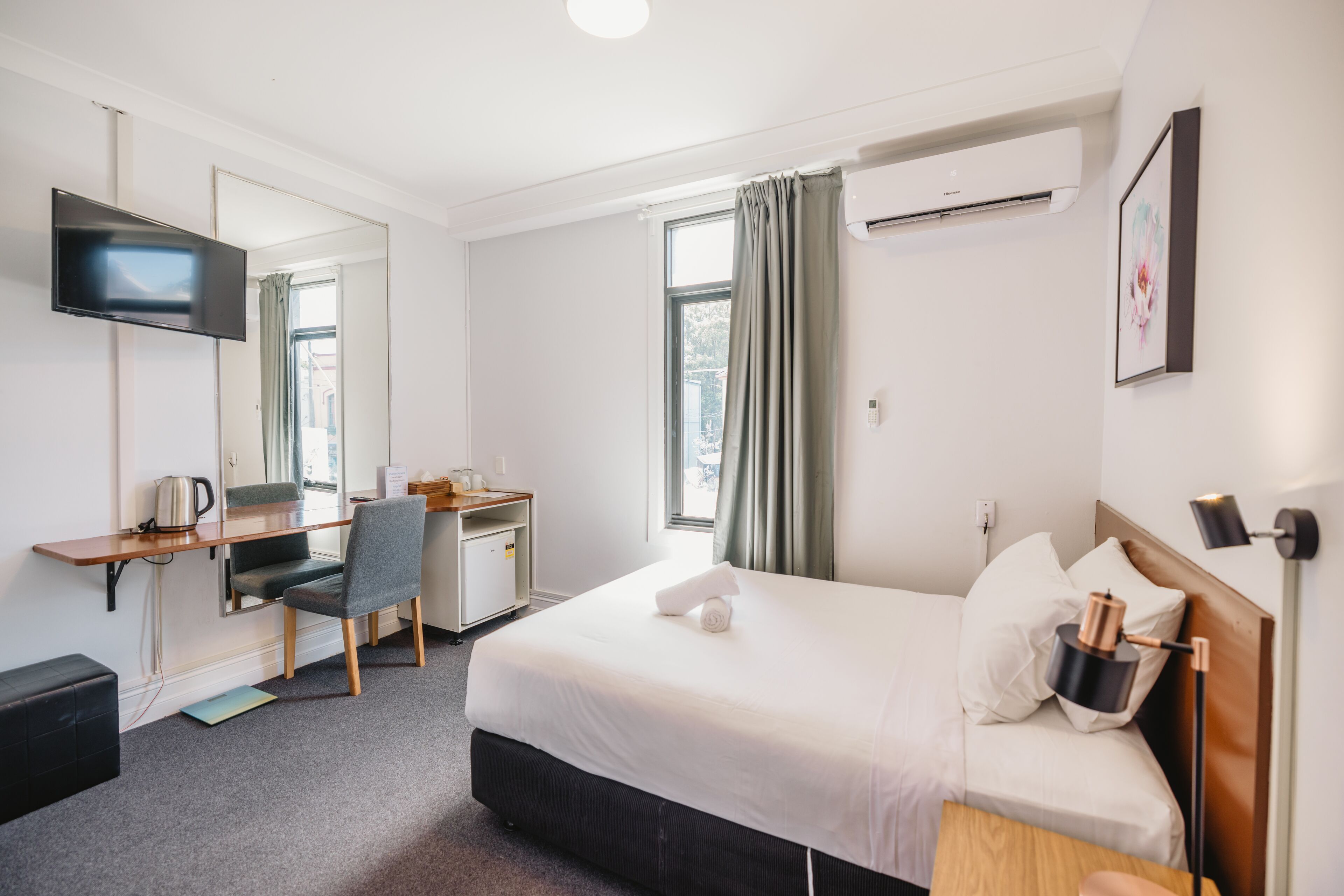 Una habitación de hotel con cama, escritorio, silla, televisión, aire acondicionado y ventana con cortinas.