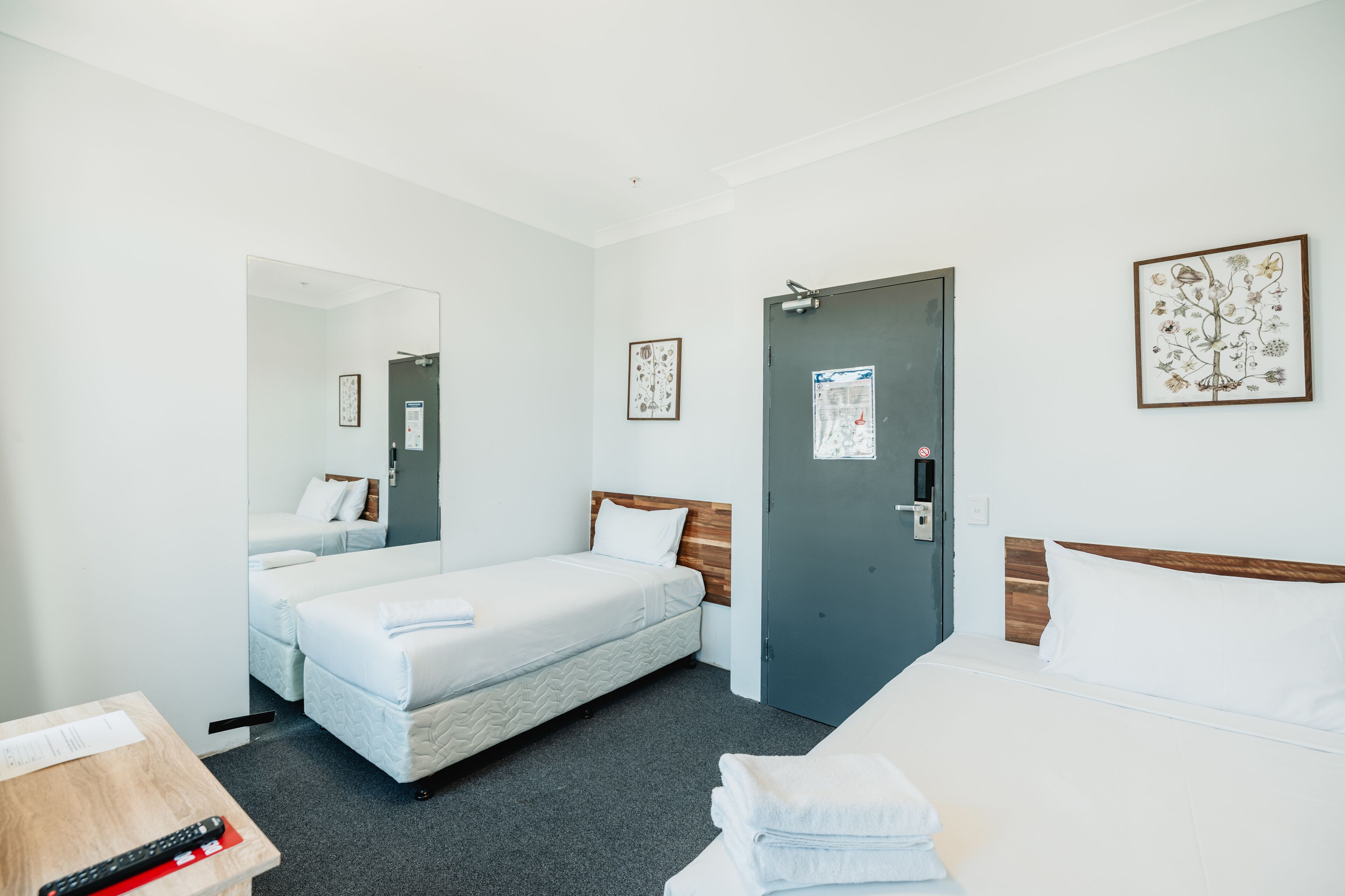 A hotel room with two single beds, a wooden headboard, a grey door, and a small table with a remote control.