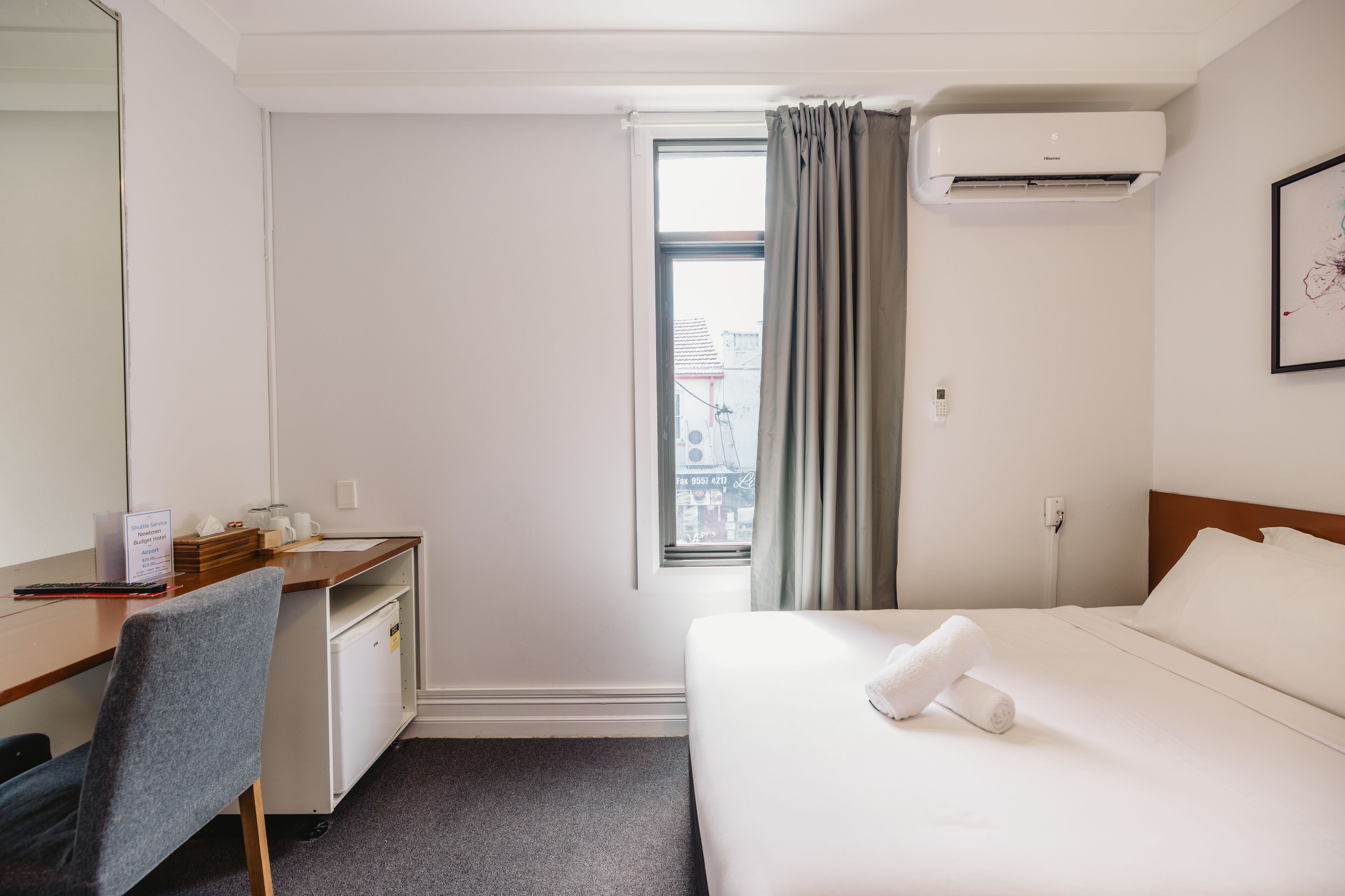 Une chambre d’hôtel comprenant un lit, un bureau, une chaise, une fenêtre avec des rideaux, un climatiseur et un tableau encadré au mur.