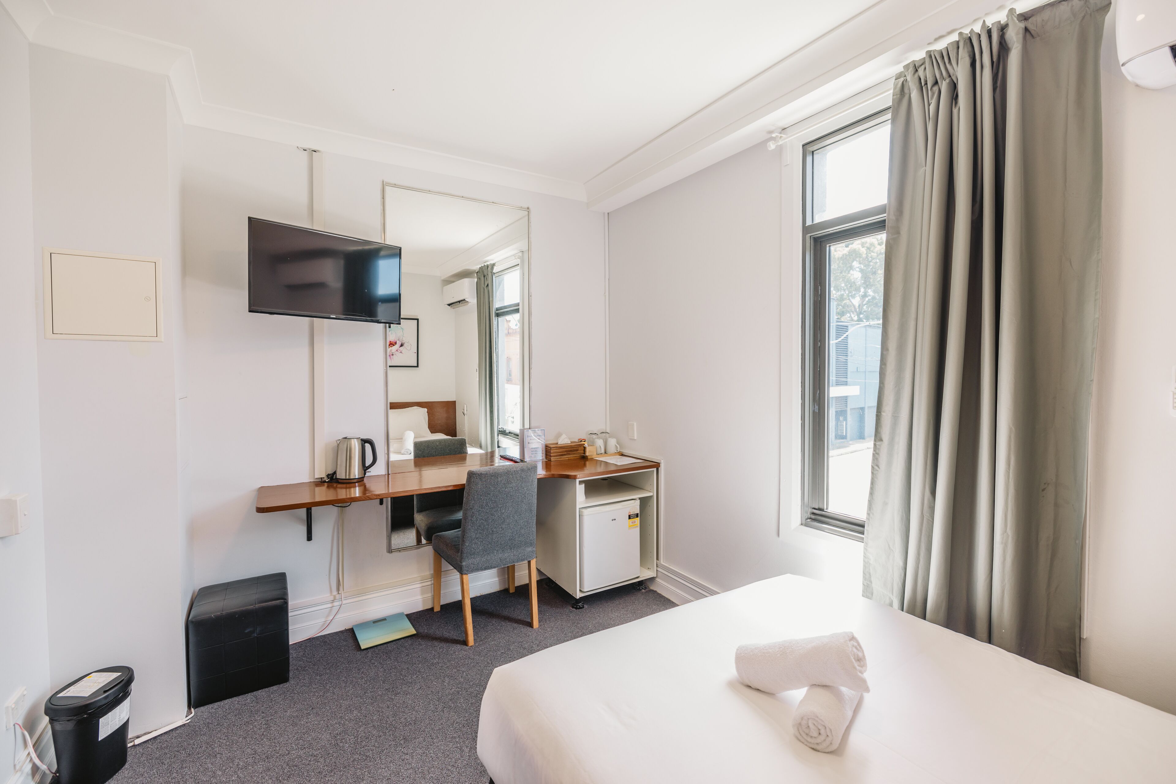 Ein Hotelzimmer mit Bett, Schreibtisch, Stuhl, Fernseher, einem Fenster mit Vorhängen und einer kleinen Küchenzeile.